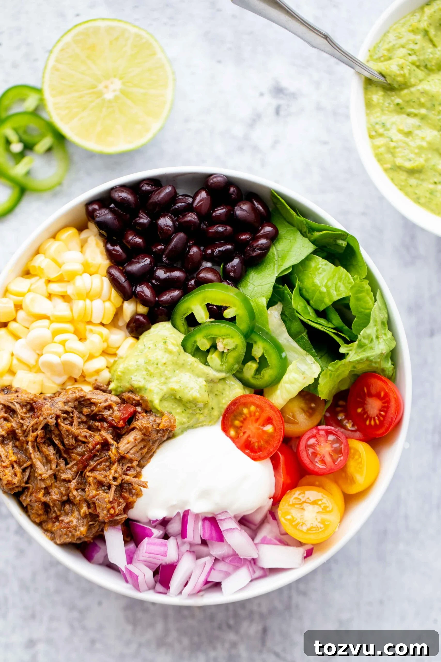 Tender Instant Pot Shredded Beef for Tacos and Sandwiches 3 overhead photo of a burrito bowl with shredded beef corn black beans lettuce tomatoes diced red onion sour cream jalapeños and avocado sauce on a concrete background