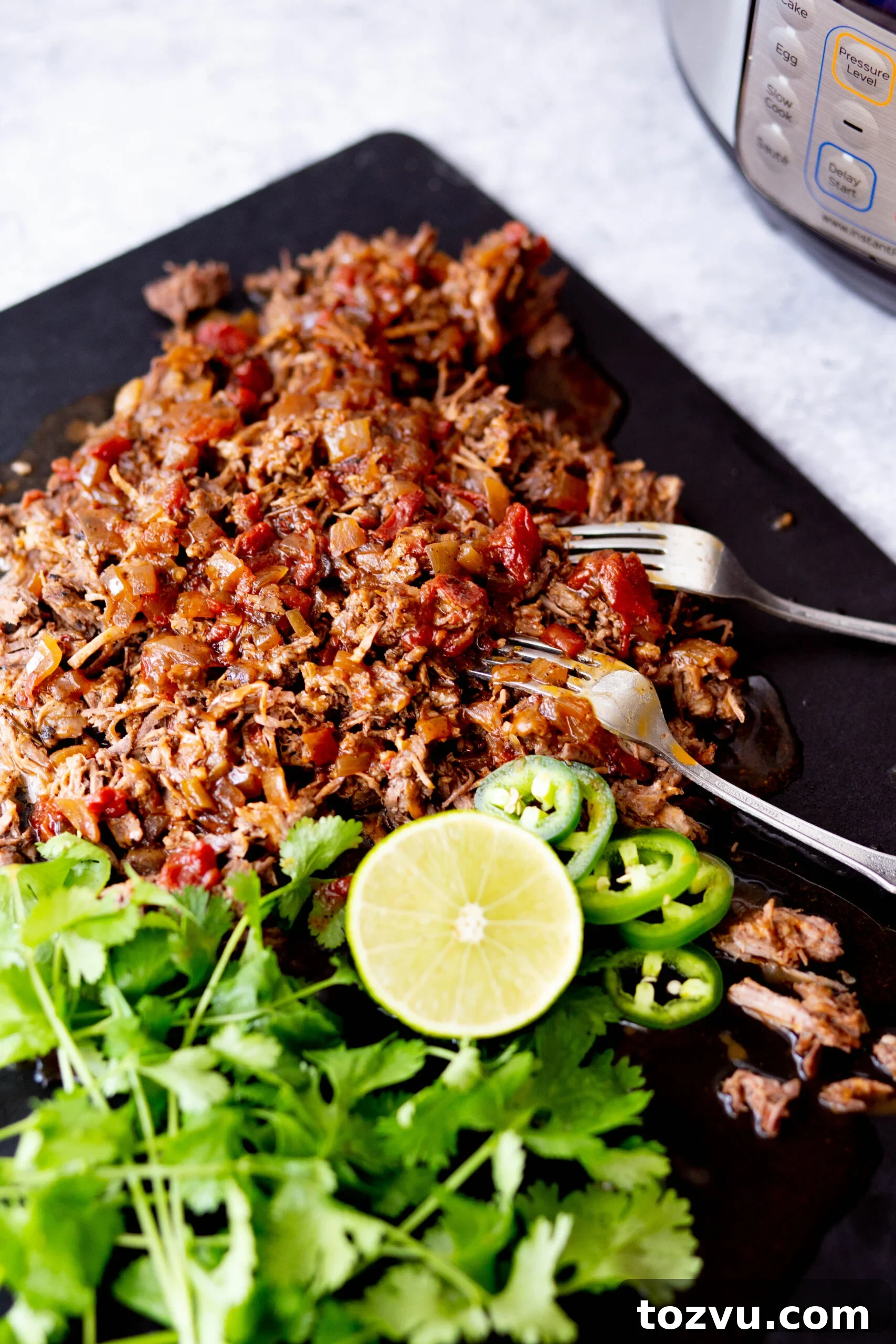 Tender Instant Pot Shredded Beef for Tacos and Sandwiches 4 45 degree photo of instant pot Mexican shredded beef on a black cutting board with cilantro lime and jalapeño and two forks and instant pot in the background