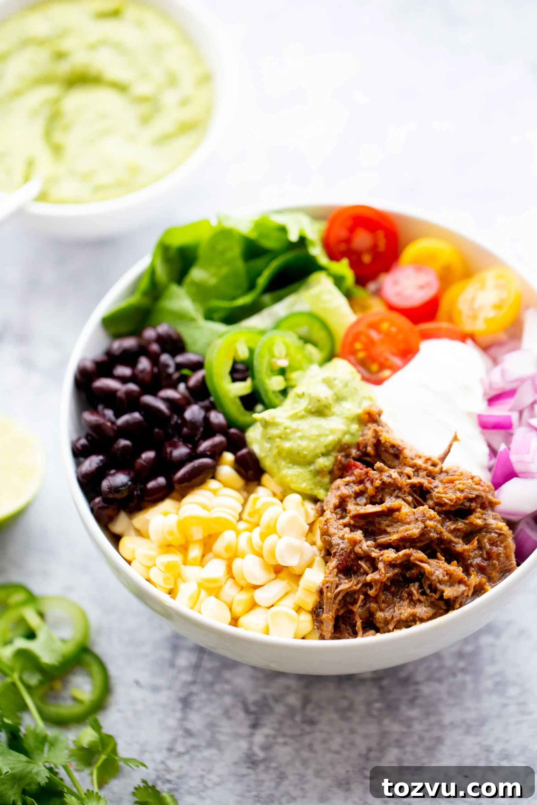 Tender Instant Pot Shredded Beef for Tacos and Sandwiches 5 45 degree photo of a backlit burrito bowl with shredded beef corn black beans lettuce tomatoes diced red onion sour cream jalapeños and avocado sauce on a concrete background with a bowl of avocado sauce in the background