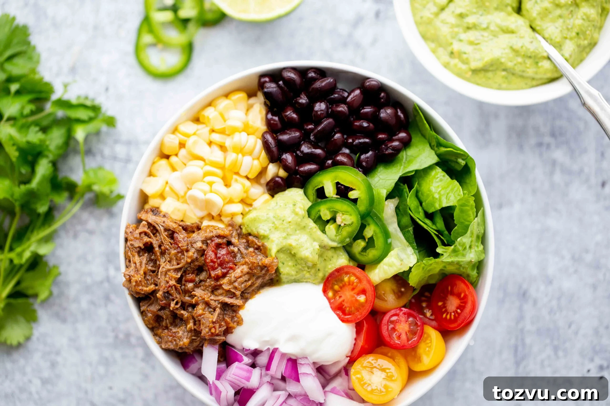 Tender Instant Pot Shredded Beef for Tacos and Sandwiches 6 closeup overhead photo of a burrito bowl with shredded beef corn black beans lettuce tomatoes diced red onion sour cream jalapeños and avocado sauce on a concrete background with a bowl of avocado sauce in the background