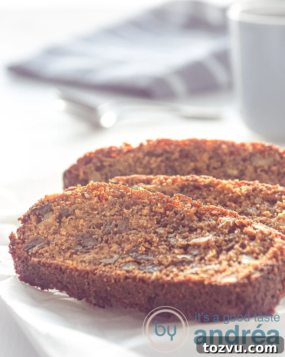 Three slices of date and apple cake on a white plate. A grey mug and cloth in the back.