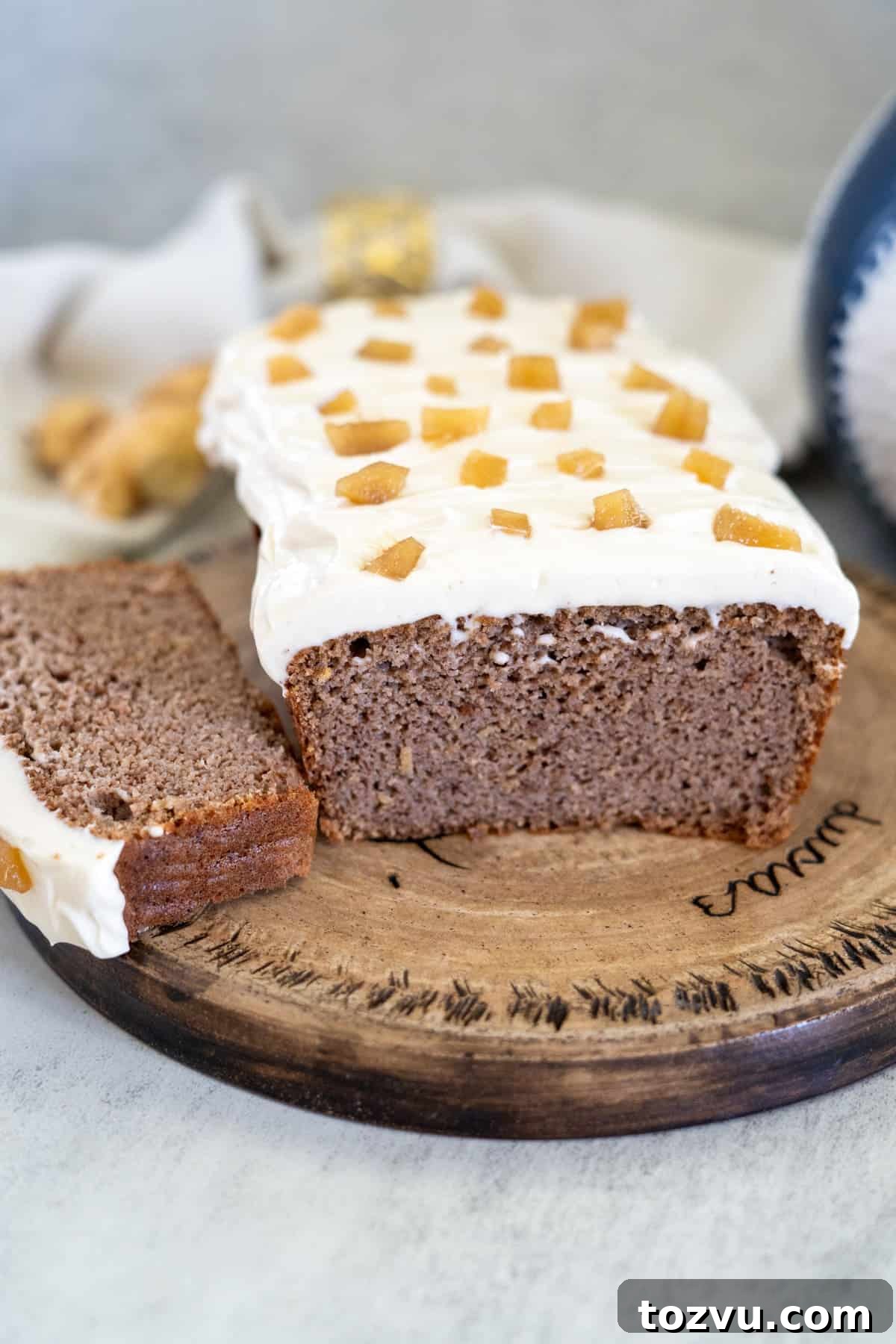 A sliced loaf of frosted ginger cake with small golden pieces on top is displayed on a round wooden board.