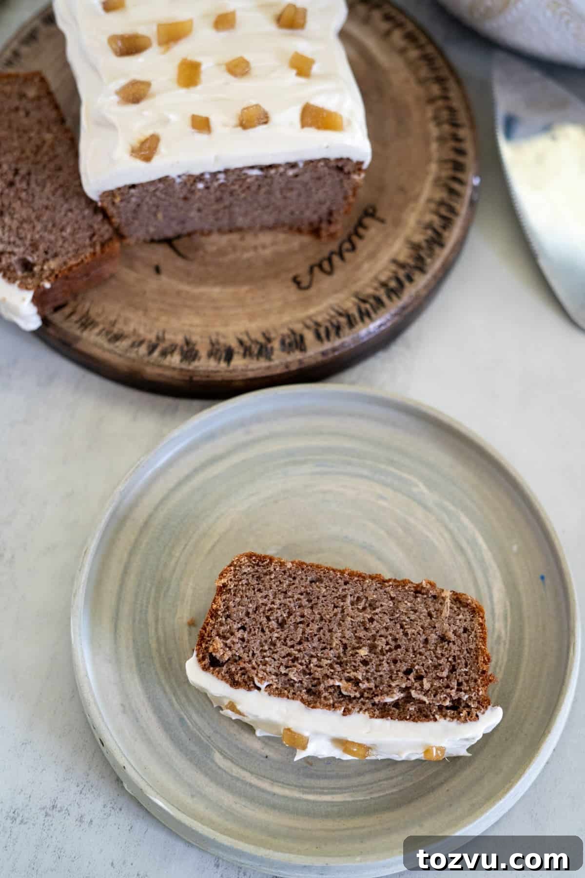 A slice of ginger cake with frosted icing sits on a plate, adorned with candied ginger pieces. In the background, the rest of the delicious cake rests on a wooden platter.