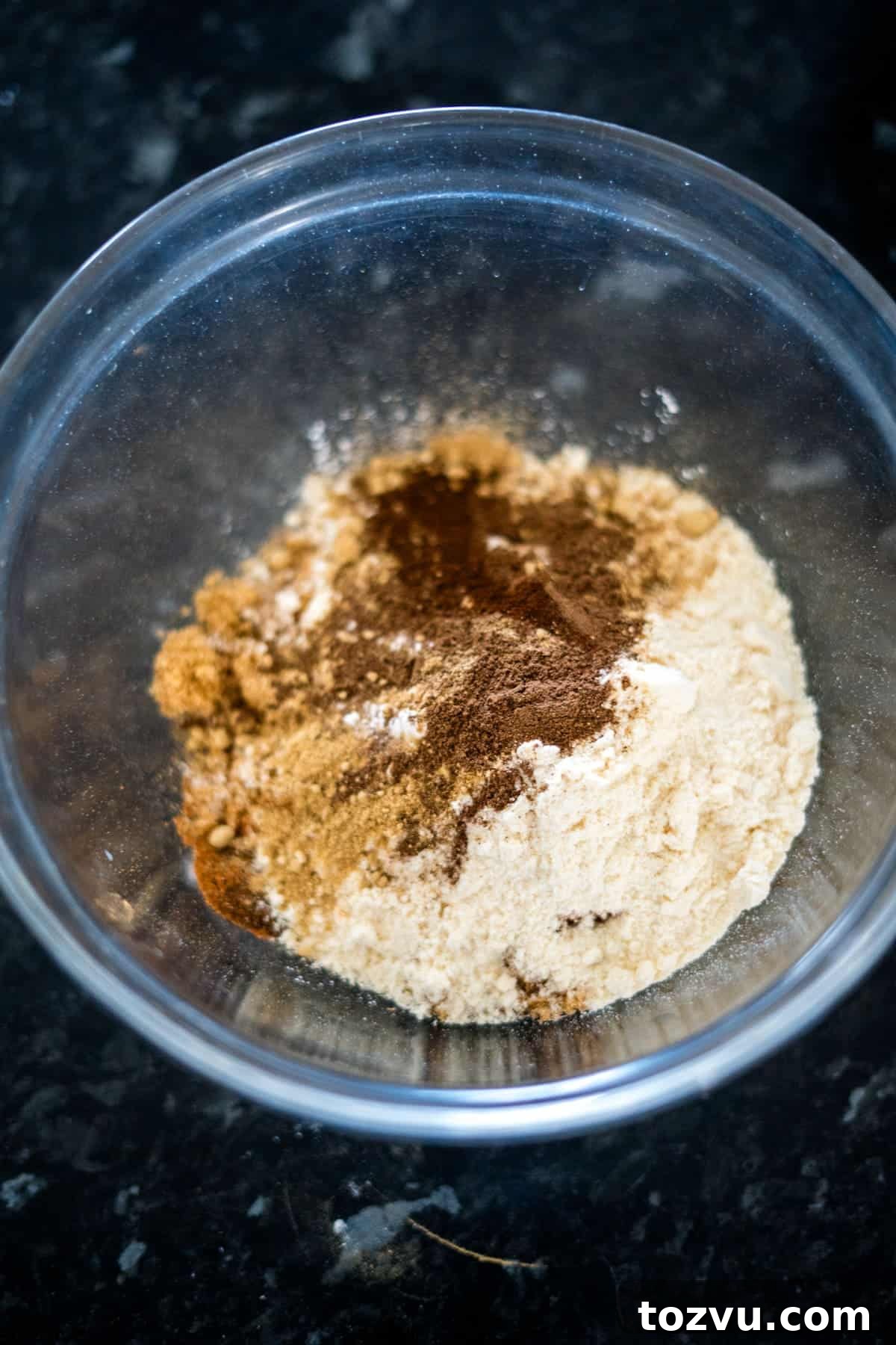 A mix of dry ingredients for a delicious ginger cake, including flour and spices, sits elegantly in a transparent glass bowl on a dark countertop.