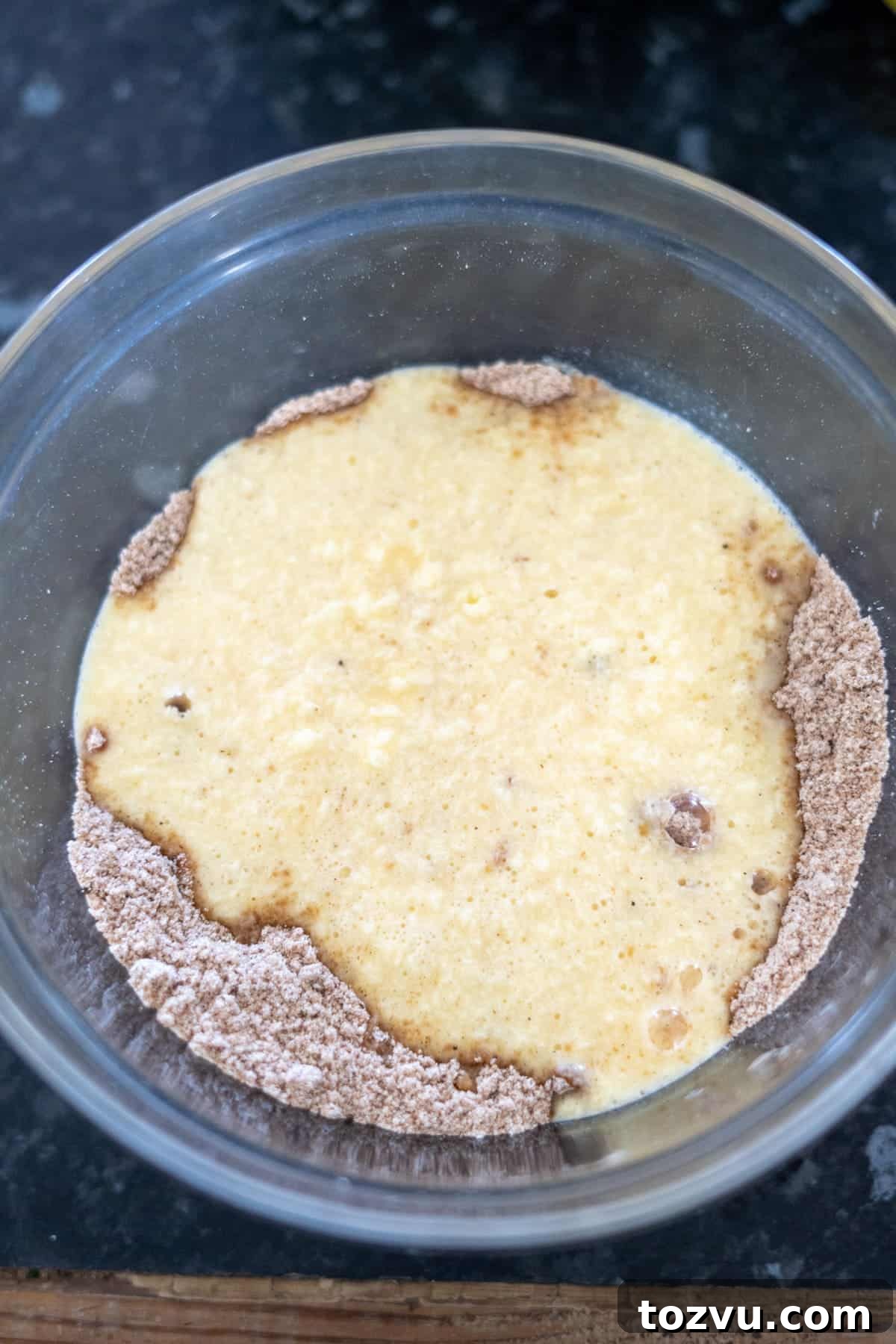 A glass bowl with a mixture of wet and dry ingredients partially combined for a ginger cake, sitting on a dark countertop.