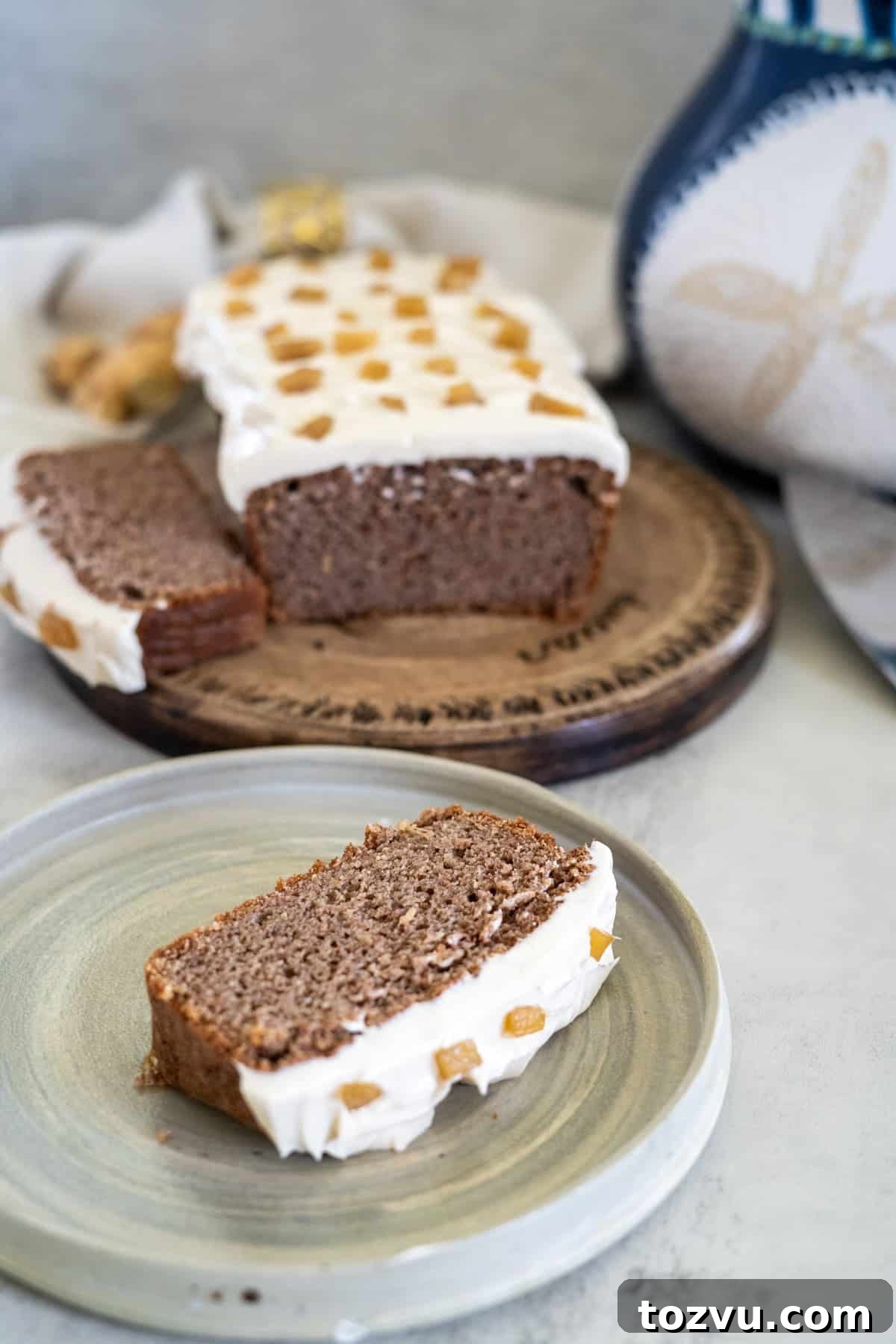 A slice of frosted ginger cake with candied ginger on top sits on a plate, in front of the rest of the loaf resting on a wooden board.
