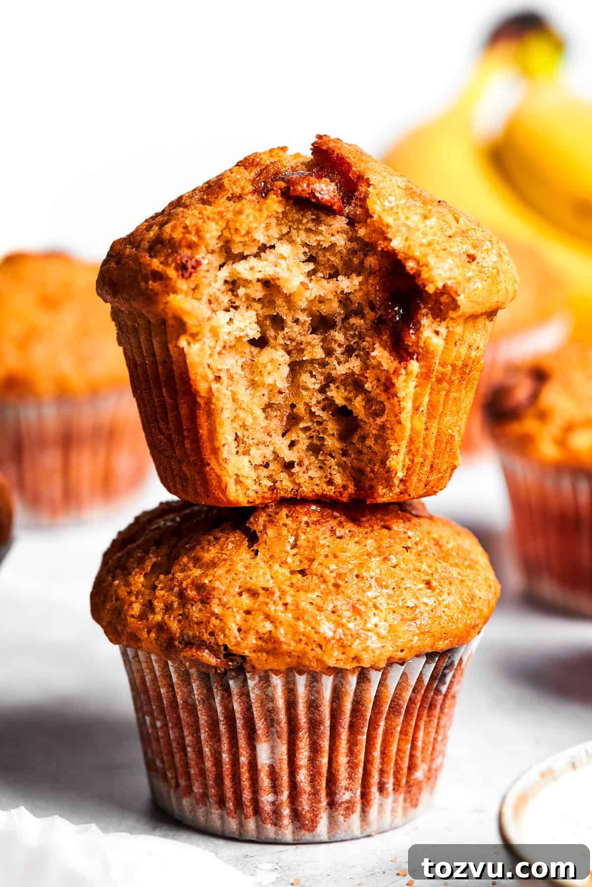 A banana muffin bitten into stands on top of a whole banana muffin.