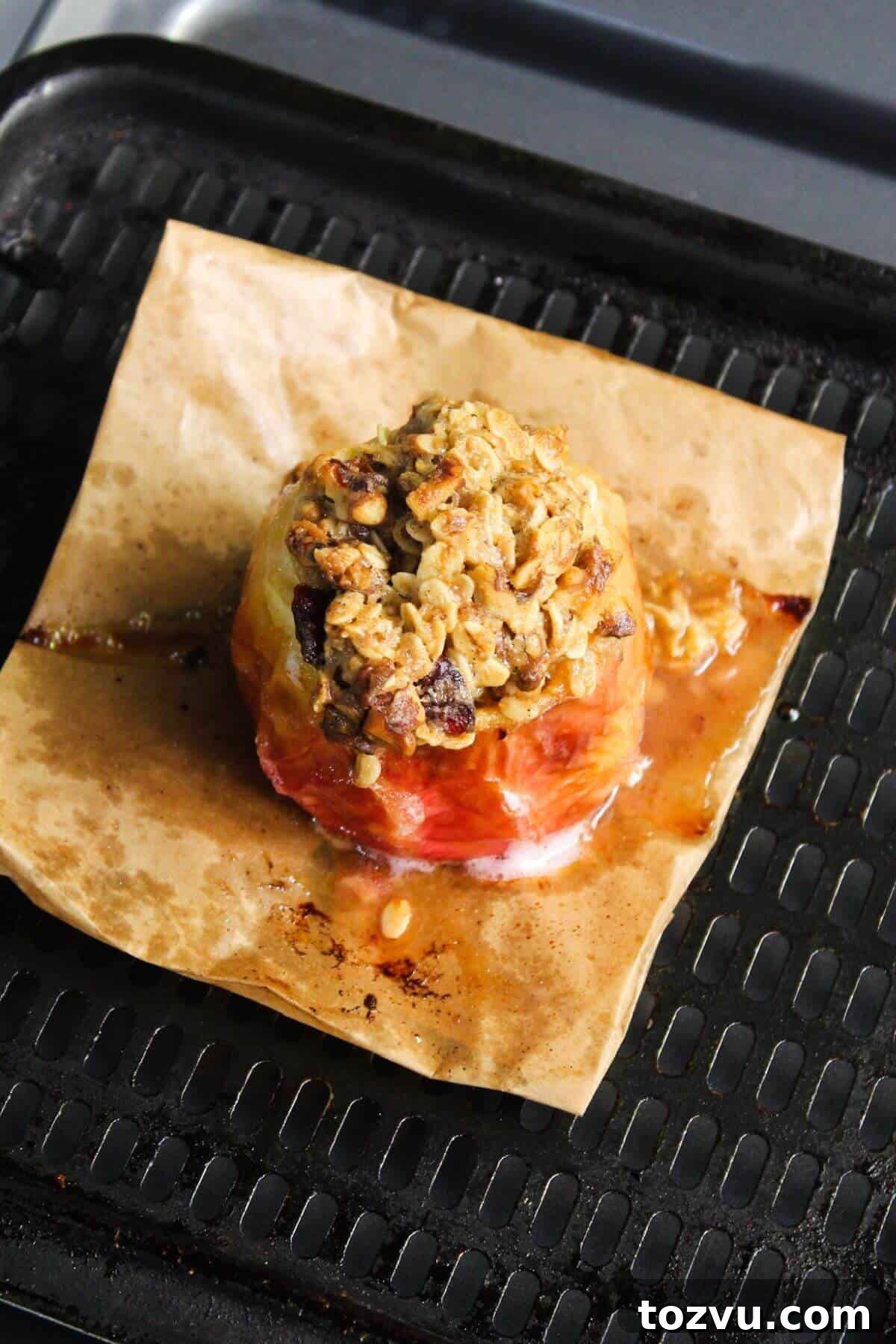 Air Fryer Baked Apples with Cinnamon and Brown Sugar 9 Baked apple with a golden oat and nut topping rests on parchment paper, nestled perfectly in a black grill pan for that crisp finish, reminiscent of an air fryer's touch.