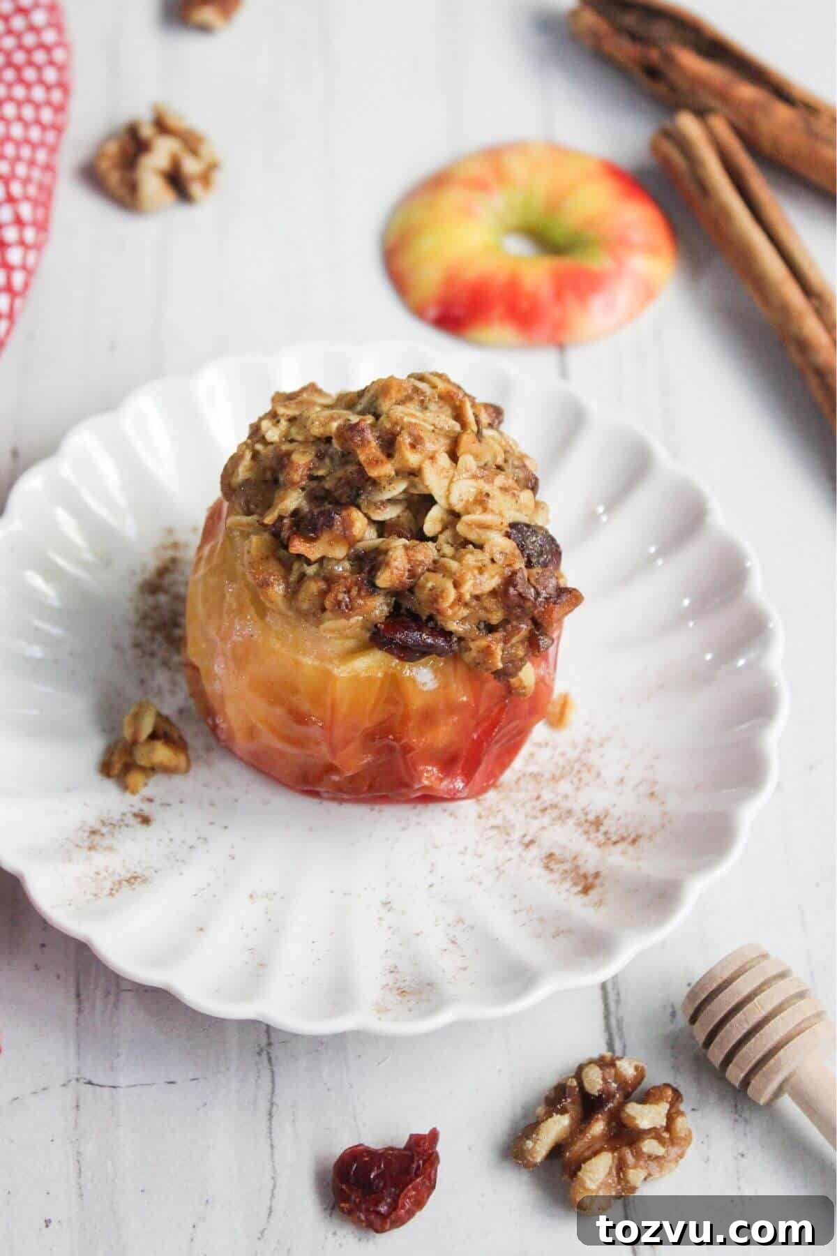 Air Fryer Baked Apples with Cinnamon and Brown Sugar 10 Baked apple in an air fryer topped with oats and nuts on a white plate, surrounded by walnuts, cinnamon sticks, an apple slice, and a honey dipper.