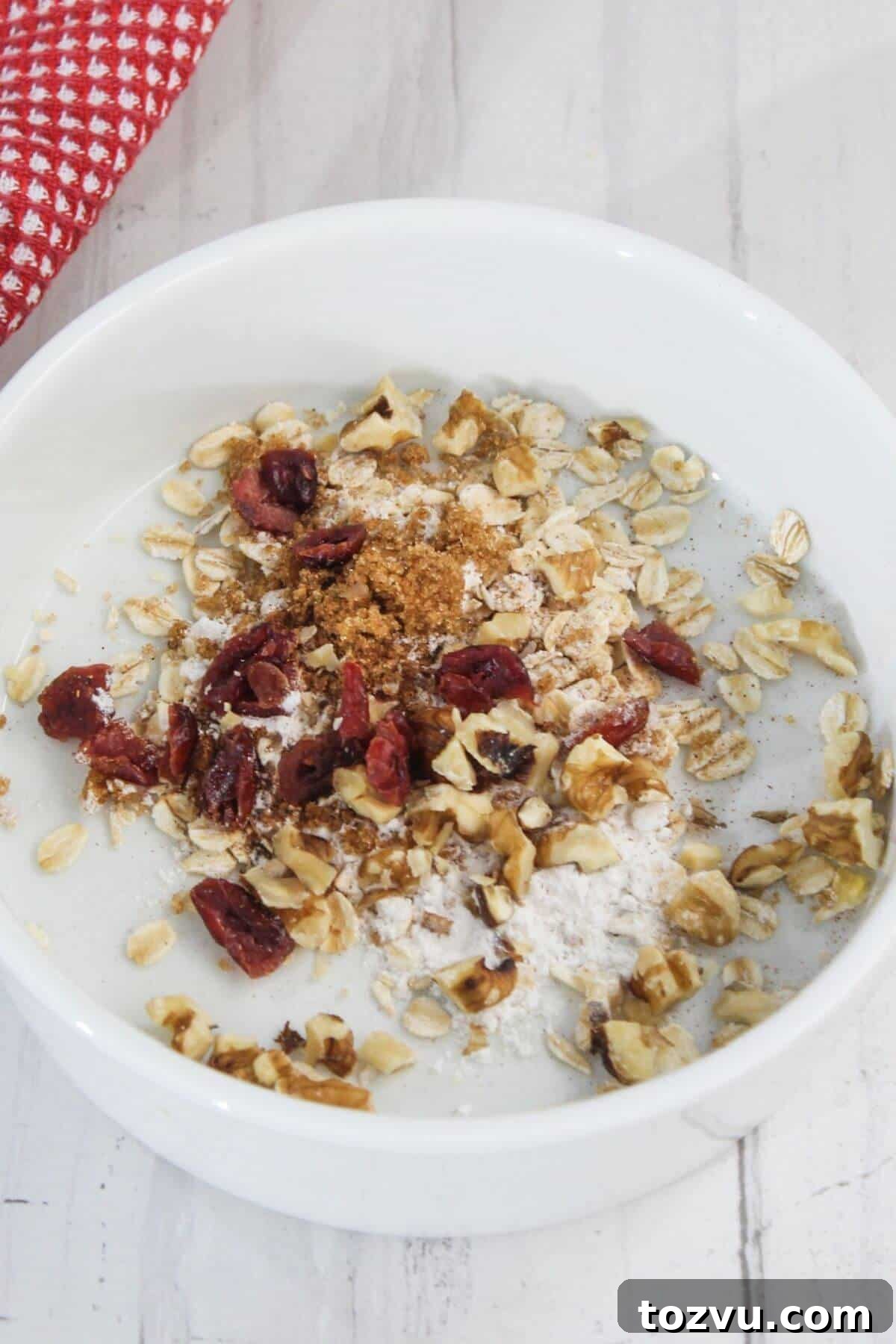 Air Fryer Baked Apples with Cinnamon and Brown Sugar 4 A white bowl holds oats, dried cranberries, walnuts, cinnamon, and brown sugar. Nestled beside it on a light wooden surface with a red and white checkered cloth is a perfectly baked apple from the air fryer, adding warmth to your cozy breakfast scene.