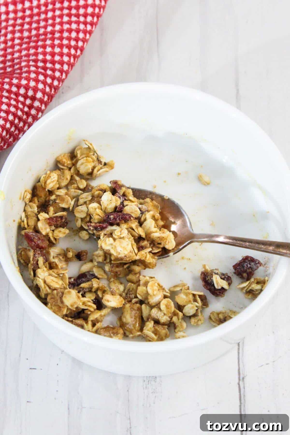Air Fryer Baked Apples with Cinnamon and Brown Sugar 5 A white bowl containing partially eaten oatmeal with a spoon inside rests on a light-colored surface. A red and white cloth is visible nearby, alongside the delightful aroma of baked apple in an air fryer wafting through the room.