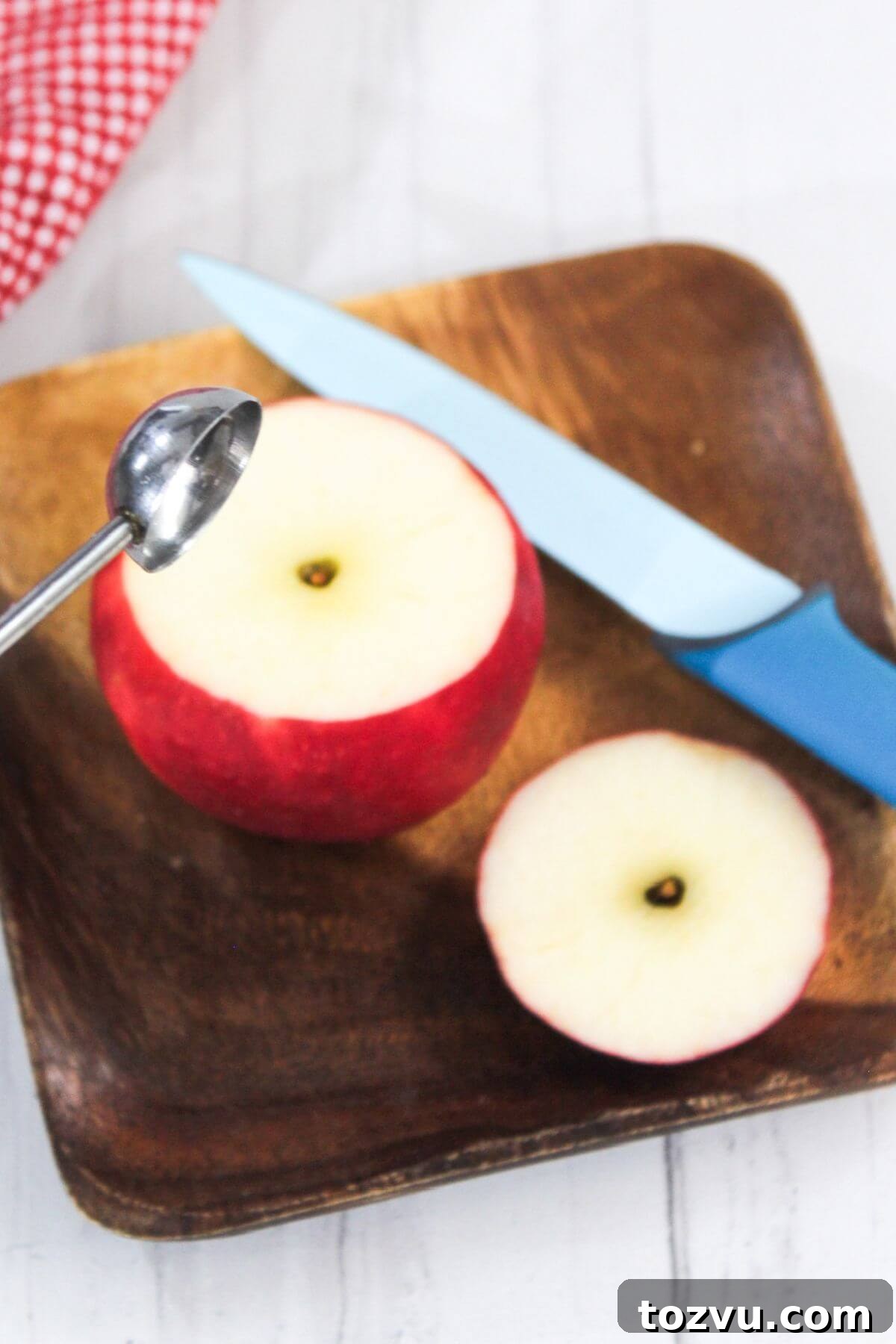 Air Fryer Baked Apples with Cinnamon and Brown Sugar 6 A halved apple on a wooden plate, with one slice removed, evokes the aroma of baked apple in an air fryer. A blue-handled knife and a metal melon baller are beside it, ready for action.
