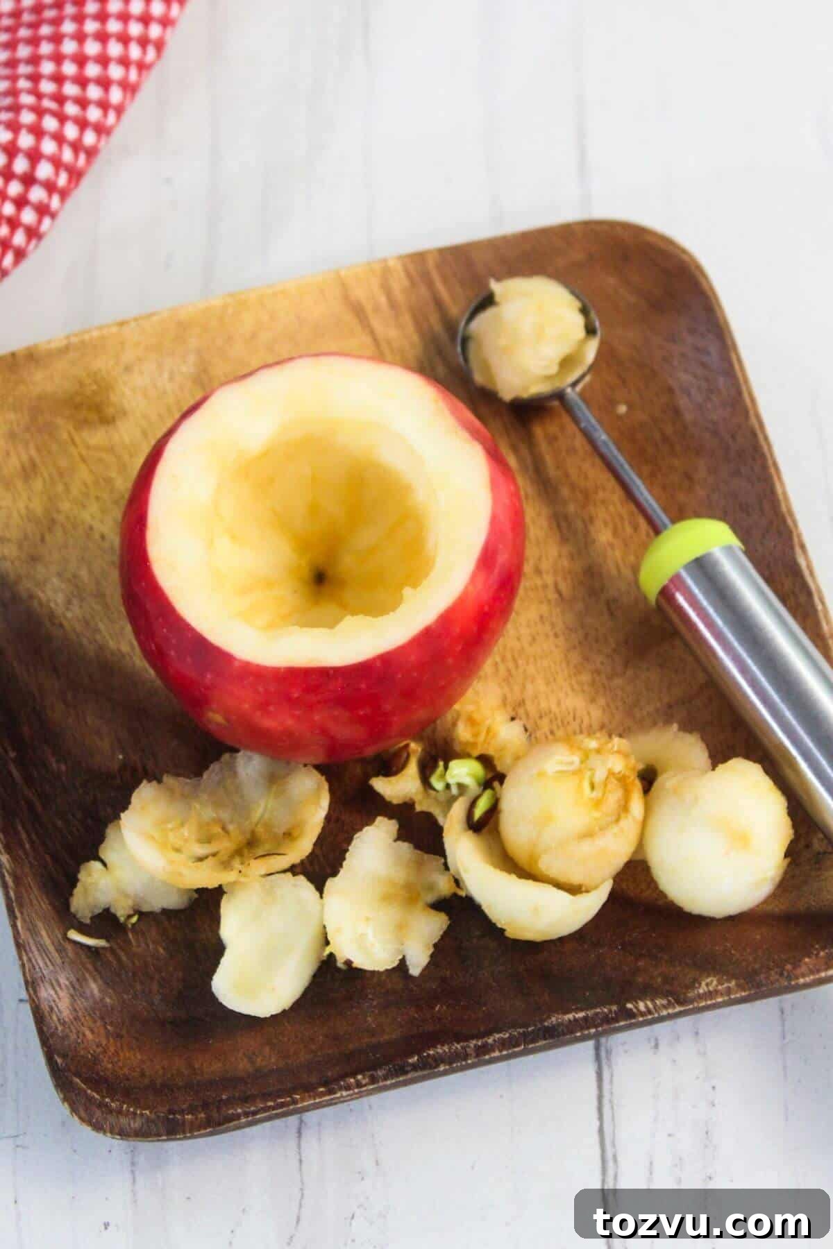 Air Fryer Baked Apples with Cinnamon and Brown Sugar 7 A cored red apple on a wooden plate, surrounded by scooped apple pieces and a melon baller, awaits transformation. Perfect for making a delicious baked apple in an air fryer.