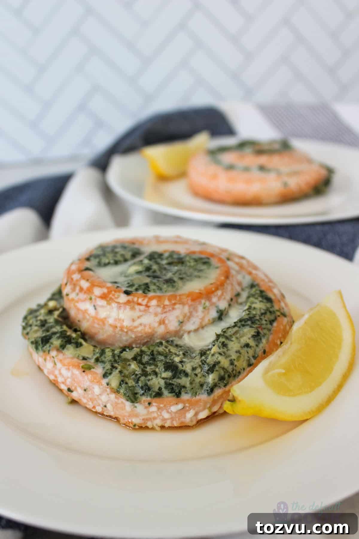 Trader Joe's Salmon Pinwheels Recipe with Feta and Spinach 2 A plate of salmon pinwheels filled with spinach and cheese, garnished with a lemon wedge. The dish is served on a white plate with a second plate of the same dish in the background, set against a chevron-patterned wall.