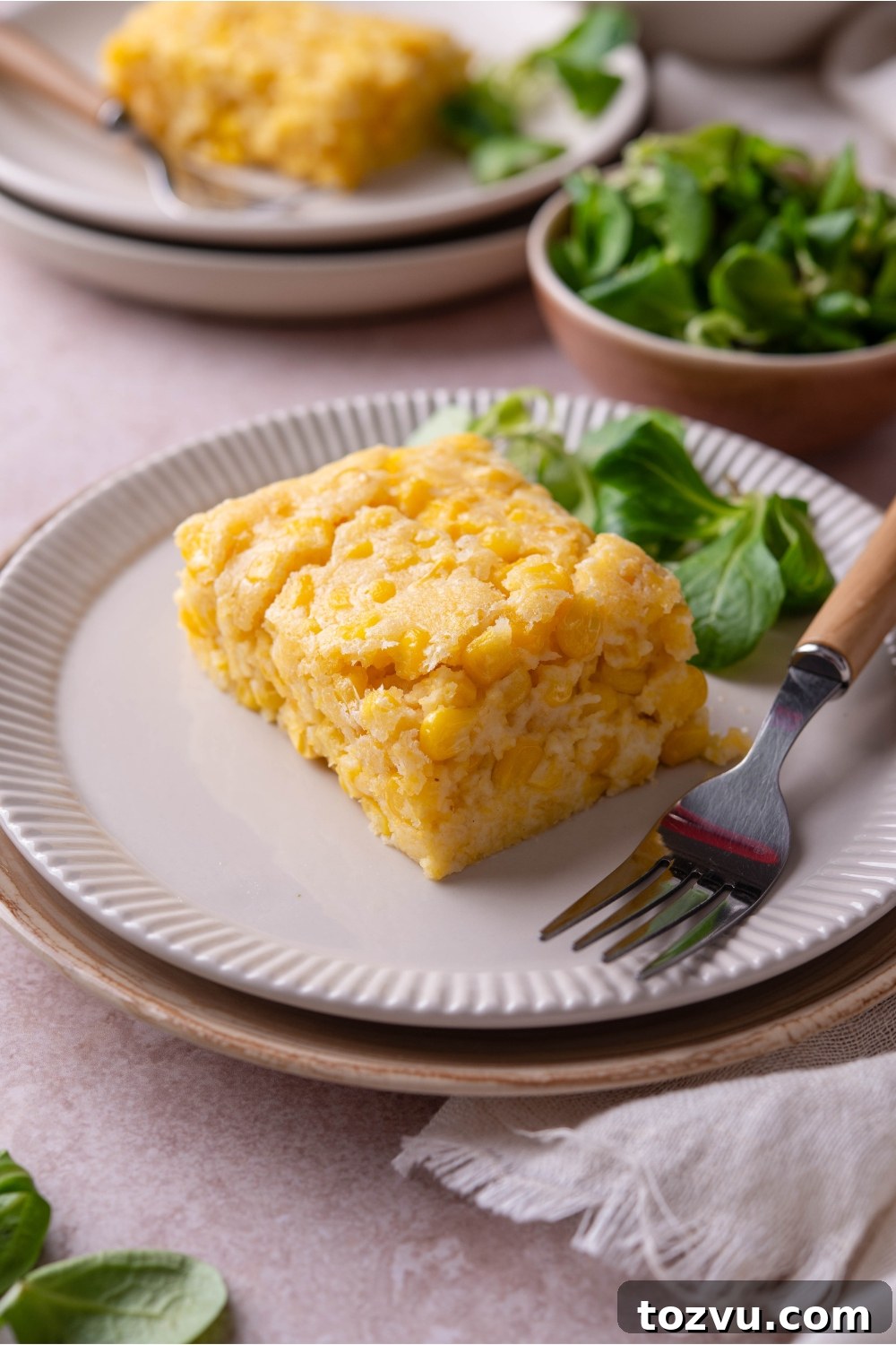 A square of corn casserole with a fork and a pile of fresh greens on the plate.