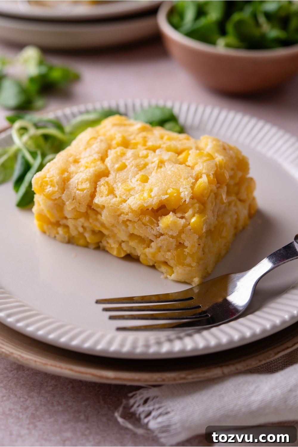 A square of corn casserole with a fork and a pile of fresh greens on the plate.