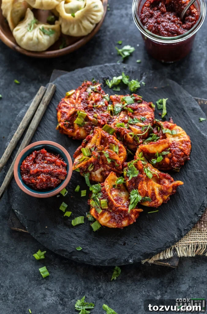 momos topped with cilantro, green onions arranged on a black board with a bowl of hot sauce and pair of chopsticks placed on the side
