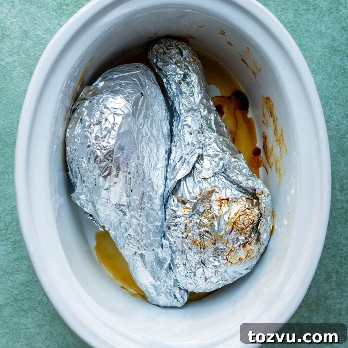 Slow-Cooker Turkey Legs with Savory Herb Glaze 3 Two foil-wrapped items placed inside a white slow cooker with some brown cooking liquid and residue visible at the bottom. The slow cooker is set on a green surface.