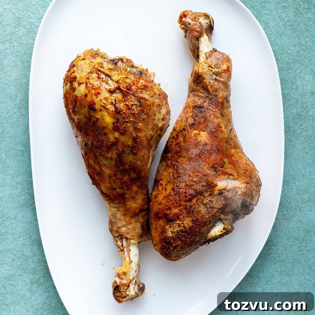 Slow-Cooker Turkey Legs with Savory Herb Glaze 4 Two seasoned and roasted turkey legs placed side by side on a white oval plate, set against a light green background.
