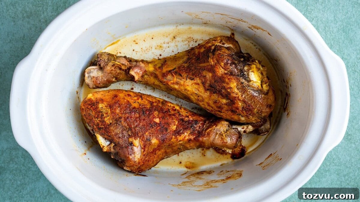 Slow-Cooker Turkey Legs with Savory Herb Glaze 9 Two seasoned and roasted turkey legs in a white oval slow cooker, with juices and browned spices visible on the bottom of the cooker. The background is a textured light green surface.