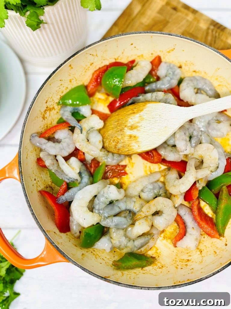 20-Minute Shrimp Fajitas with Peppers and Lime 6 raw king prawns being added into a skillet pan with red and greed diced peppers. Everything being stirred with a wooden spoon.