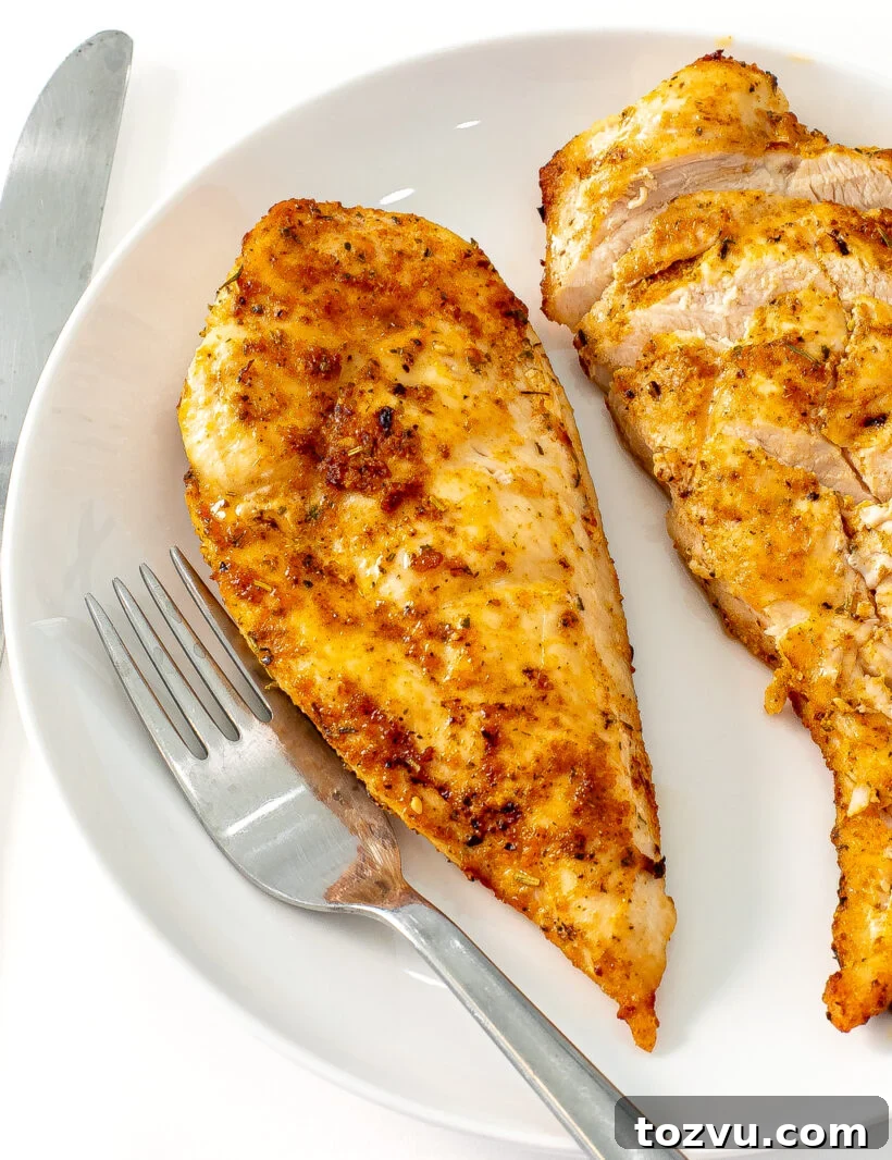 Juicy Air Fryer Chicken Breast Recipe 2 Two chicken breasts on a plate, one is sliced and the other is not, with a fork next to them.