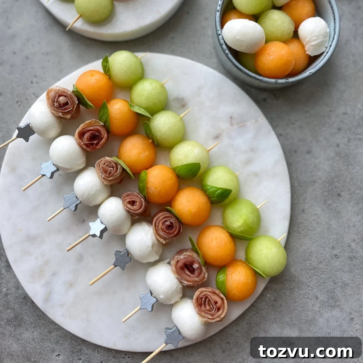 Wooden skewers with mozzarella balls, prosciutto, cantaloupe, and honeydew melon bites are garnished with fresh basil and arranged on a round marble platter.