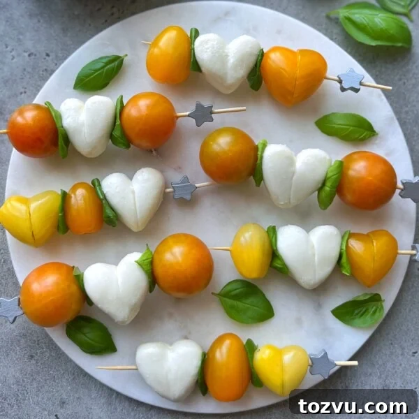 Skewers with yellow and orange cherry tomatoes, heart-shaped mozzarella, and fresh basil leaves arranged on a round marble platter.