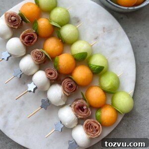 Skewers with mozzarella balls, rolled prosciutto, green and orange melon balls, and fresh basil leaves, arranged neatly on a round marble platter—perfect for healthy bites or inspiring new food recipes.