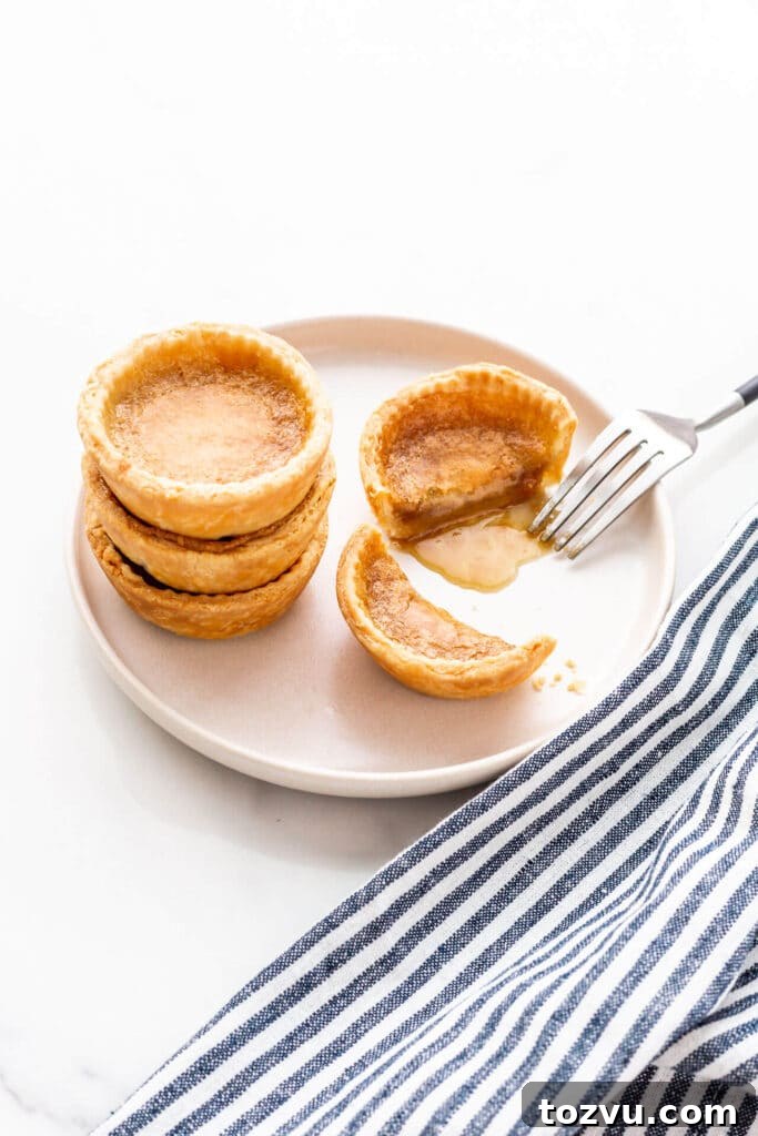 Butter Tarts Without Corn Syrup with Flaky All-Butter Crust 2 A beige plate with a stack of three butter tarts and a fourth next to it, cut open with a fork to reveal the gooey filling.