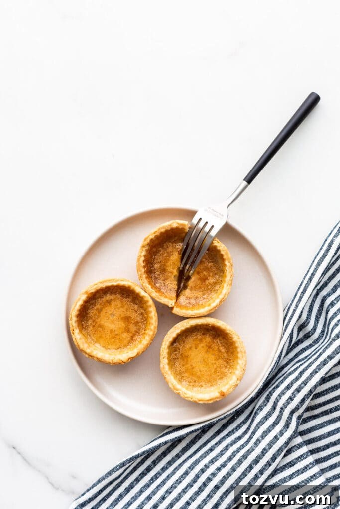 Butter Tarts Without Corn Syrup with Flaky All-Butter Crust 9 A beige plate with three butter tarts on it and a fork cutting open one of them.