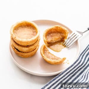 Butter Tarts Without Corn Syrup with Flaky All-Butter Crust 10 Butter tarts stacked on a beige plate with one cut open with a fork to reveal the gooey filling.