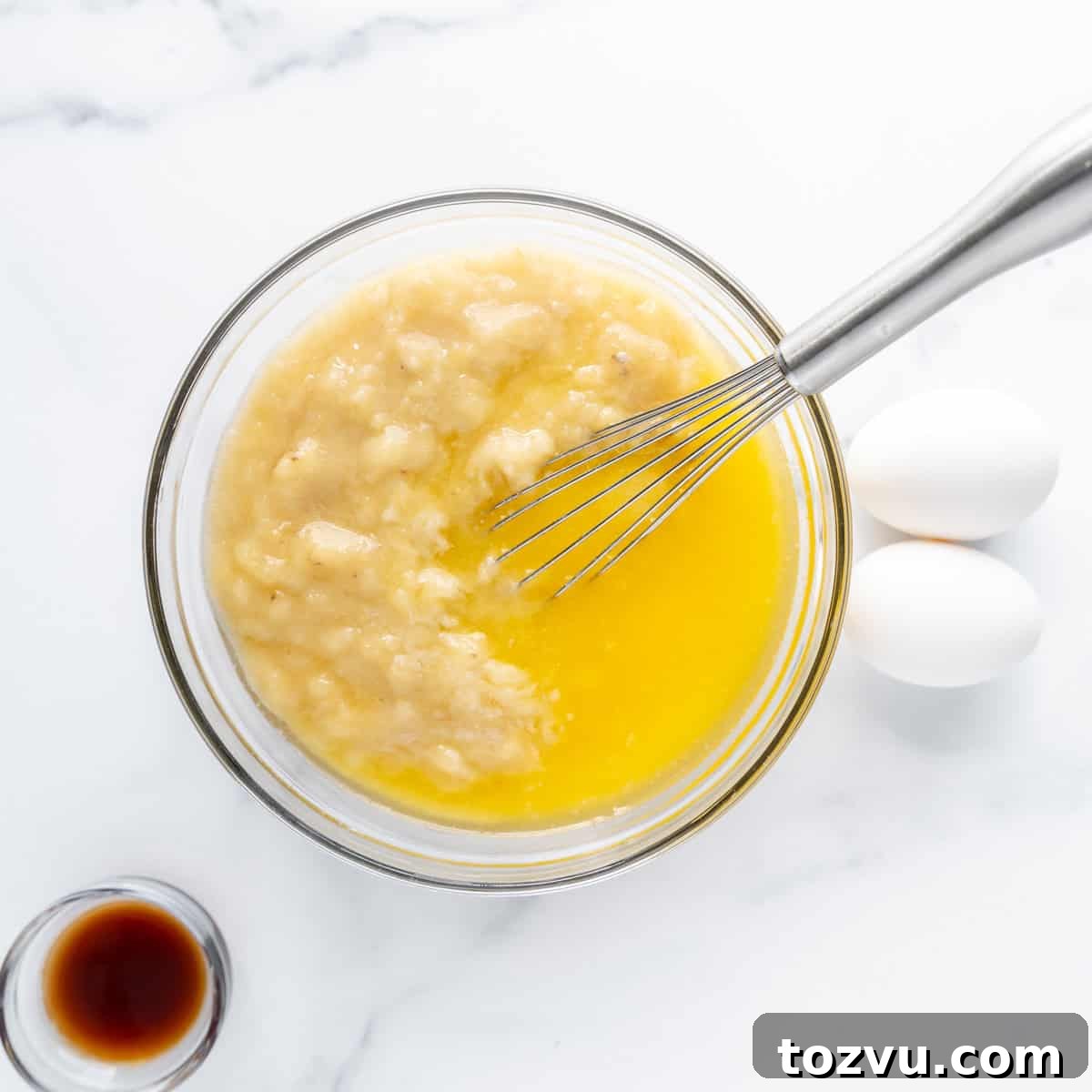 Wet ingredients in a clear bowl with a whisk. Vanilla and eggs on the side.