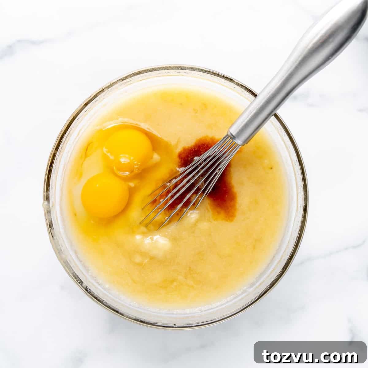 Eggs and vanilla added to wet ingredients in a clear bowl with a whisk.